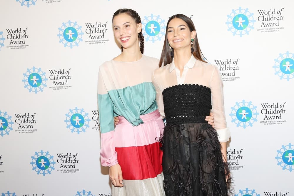 Ruby Aldridge and Lily Aldridge Followill pose for a picture in front of the step and repeat banner. The step and repeat banner has a white background with the World of Children Award logo repeated.