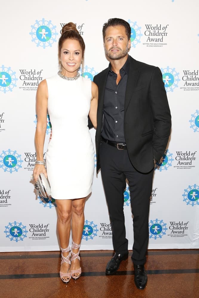 Actress Brooke Burke and singer David Charvet pose for a picture in front of the step and repeat banner. The step and repeat banner has a white background with the World of Children Award logo repeated.