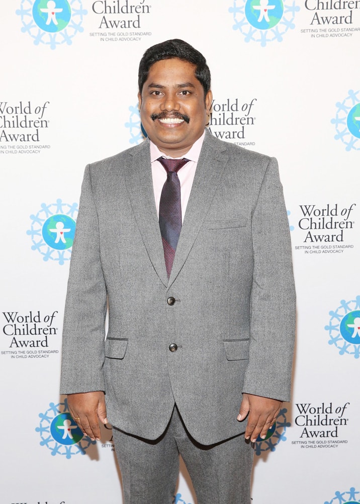 Iyyappan Subramaniyan pose for a picture in front of the step and repeat banner. The step and repeat banner has a white background with the World of Children Award logo repeated.