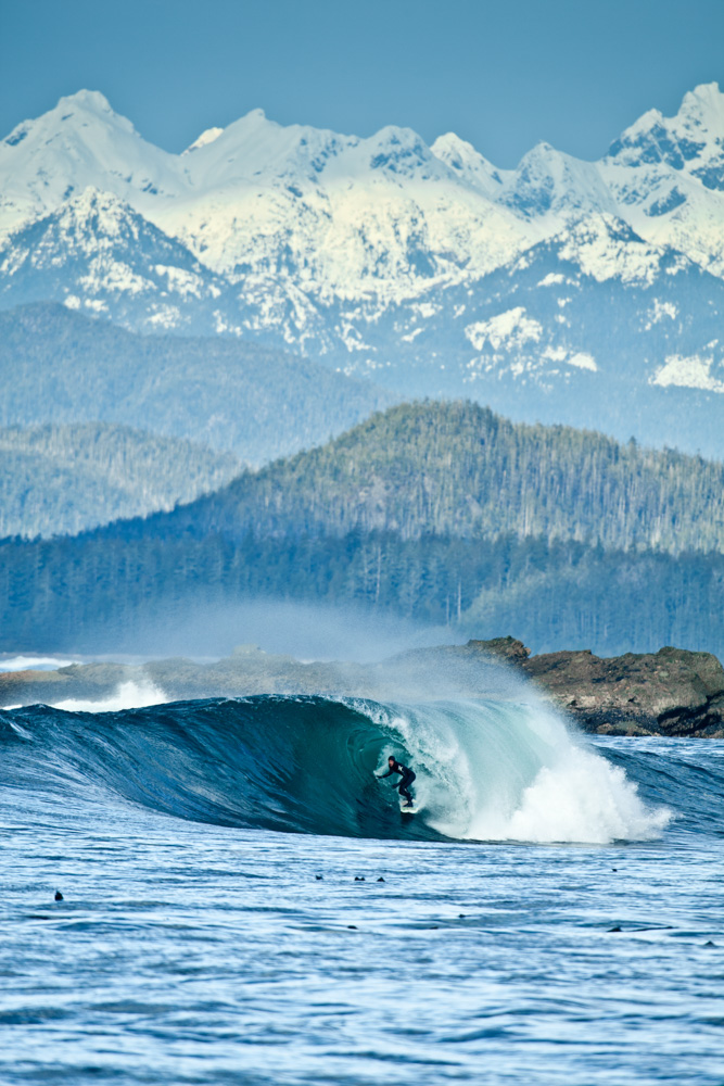 Surfer riding curled wave with greenery and snowy mountains in the background at Vancouver Island, Canada