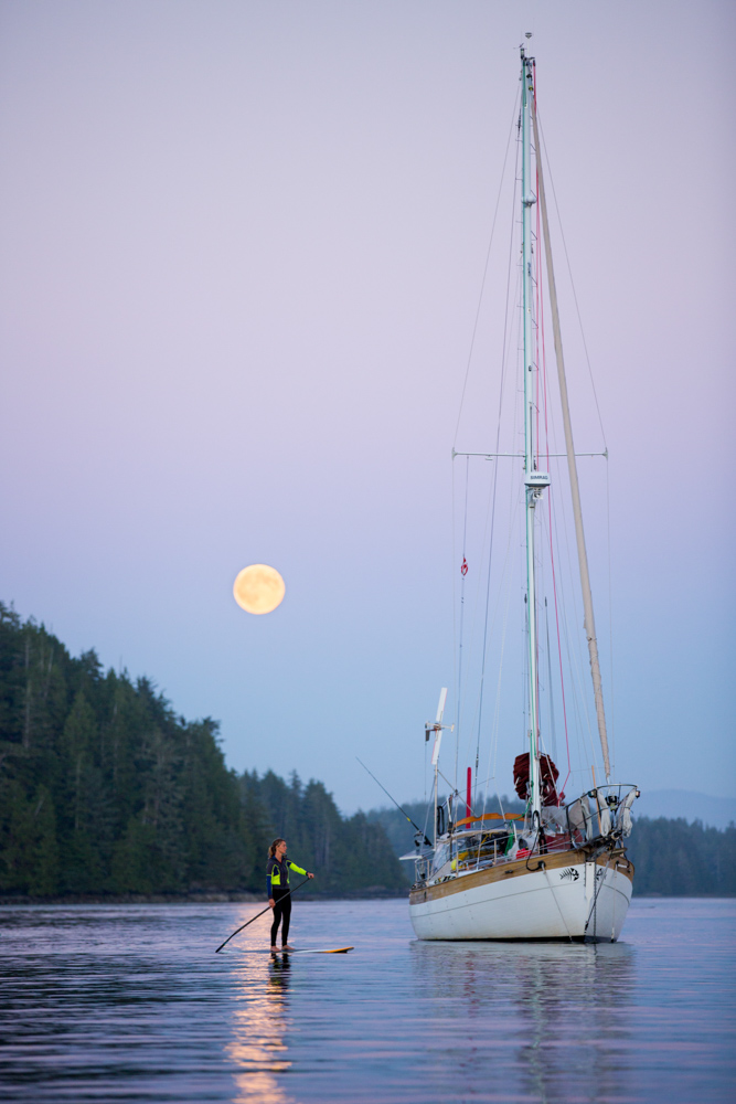 Man paddle boarding next to sailboat at twilight, with moon in the background at Vancouver Island, Canada