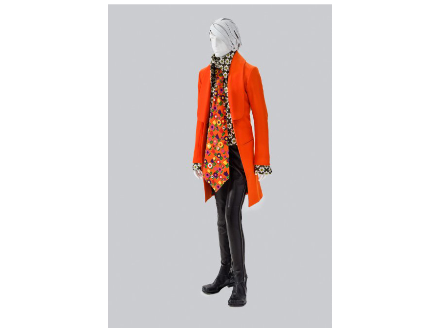 A Male Mannequin Dressed In A Bright Orange Colored Long Jacket and Patterned Tie With Leather Pants By Walter Van Beirendonck
