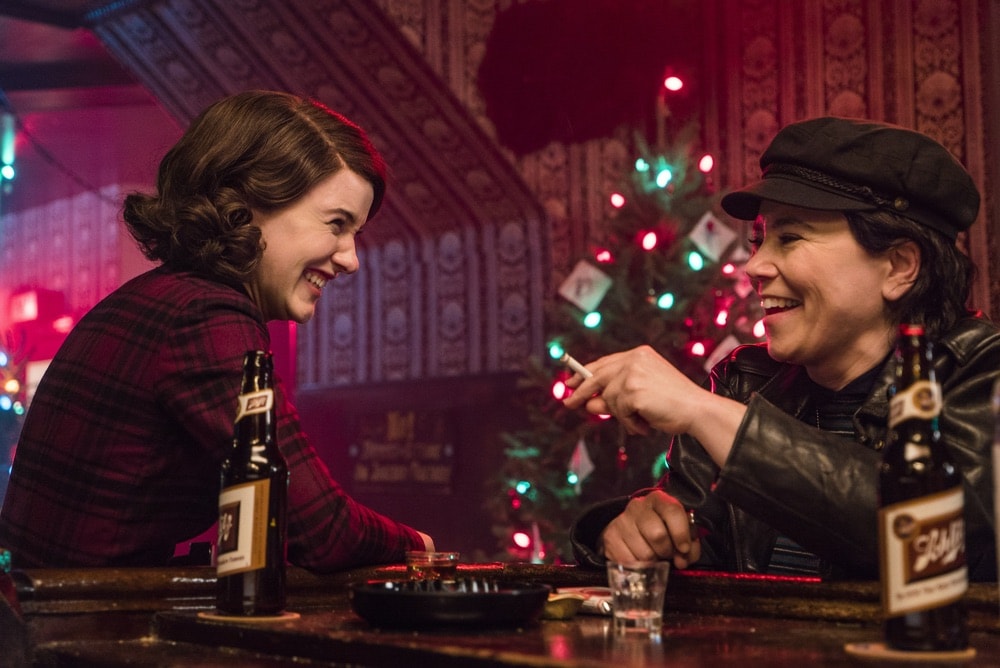 Rachel Brosnahan and Alex Borstein in Season 1 of The Marvelous Mrs. Maisel sharing a beer each at a bar at Christmas time.