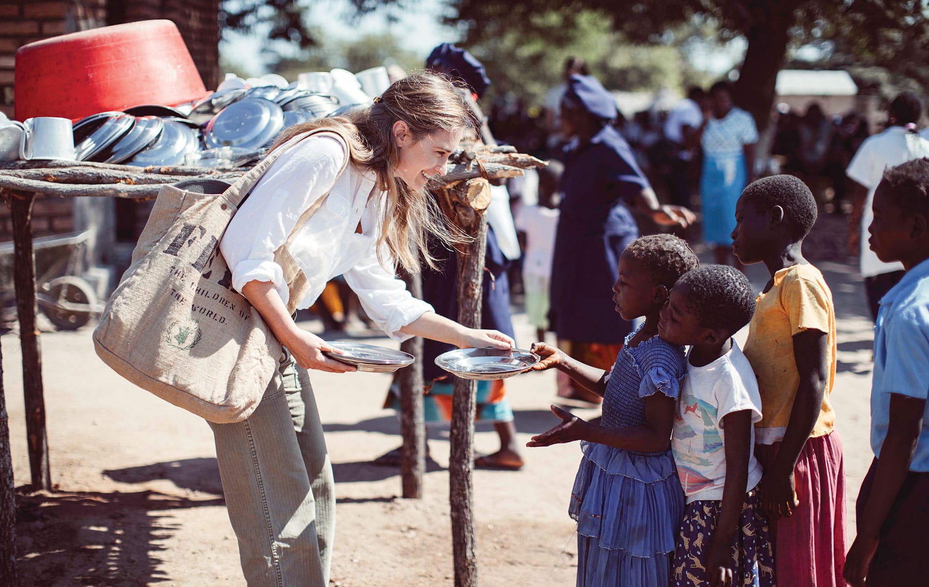 Lauren Bush Lauren handing out plates to a line of small children with a FEED bag on her shoulder