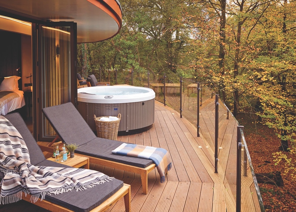 A treehouse deck containing a hot tub and 2 lounge chairs right off of the bedroom at Chewton Glen Hotel & Spa in New Forest, England surrounded by trees