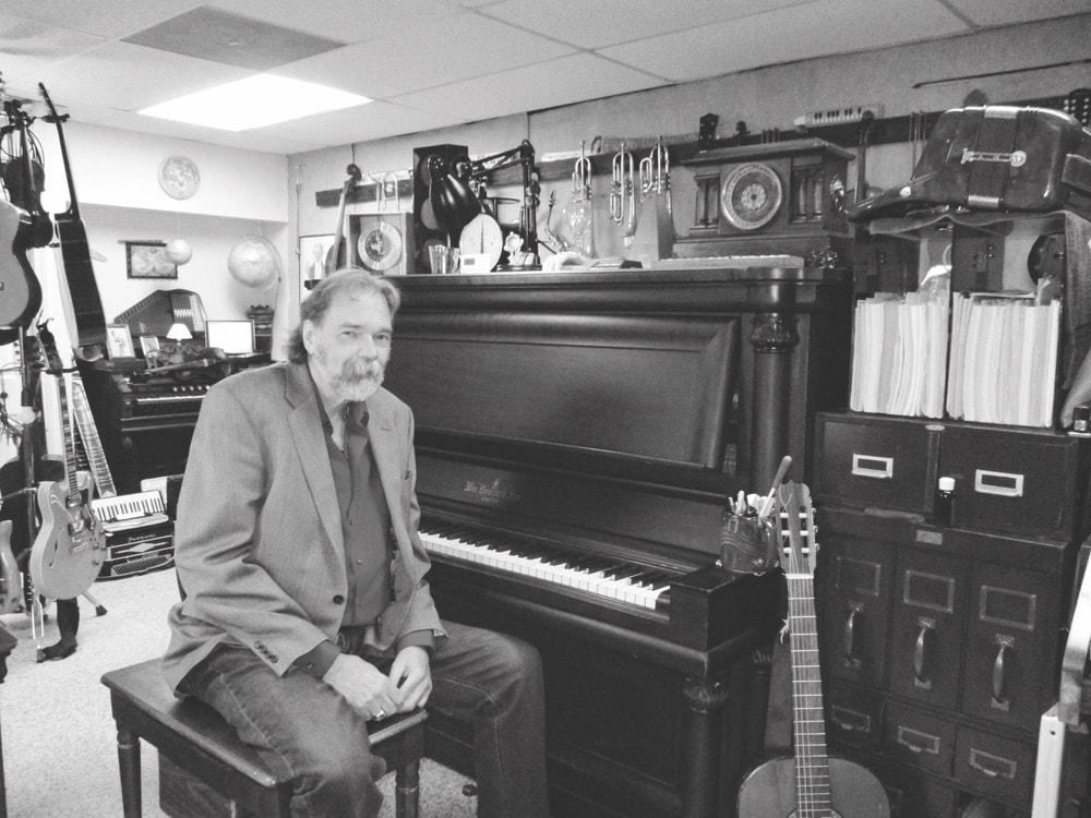 Reed sitting on a piano bench in front of his piano in black and white