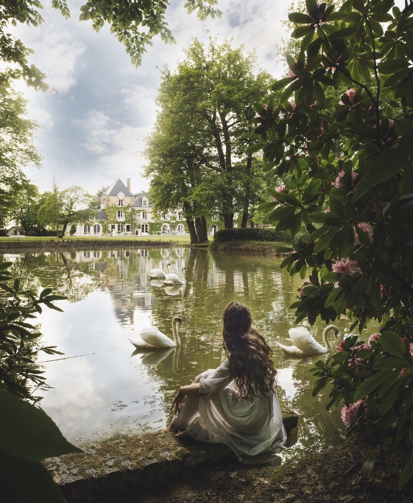 Photograph by Jamie Beck showing a female sitting on the edge of a pond surrounded by plants overlooking a large French estate and swans