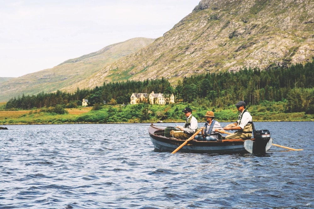 Three men fishing in one boat with a large house on the shoreline and mountains in the distance