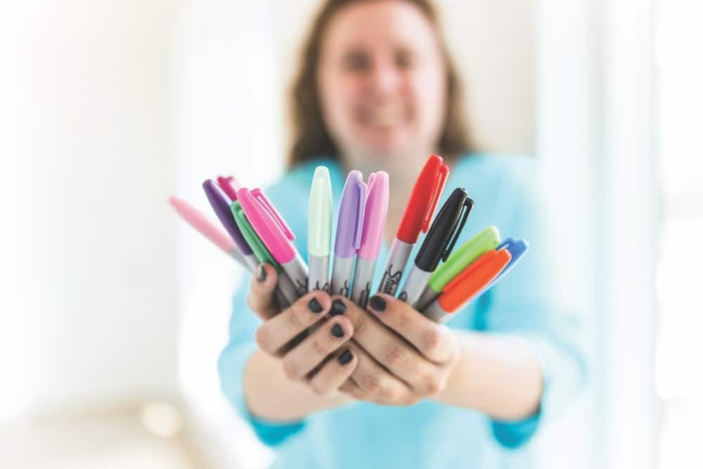 Camille Butchikas holding out a dozen colorful Sharpie markers she uses to draw cartoon-style artworks.
