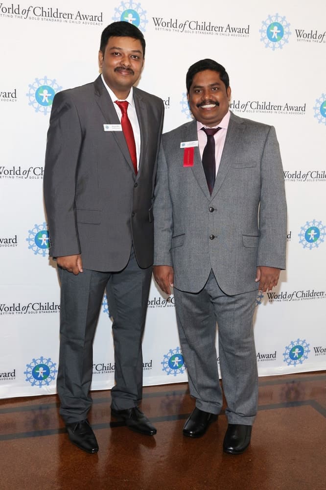 Raj Sethuraman and honoree Iyyappan Subramaniyan pose for a picture in front of the step and repeat banner. The step and repeat banner has a white background with the World of Children Award logo repeated.